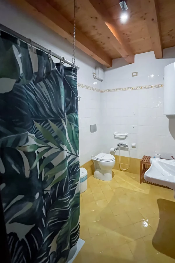 Bagno con tenda doccia a motivo foglie, pavimento giallo e soffitto con travi in legno.