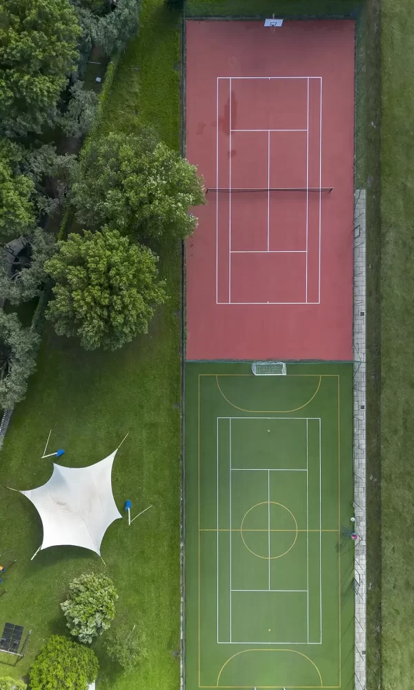 Luftaufnahme von zwei Sportplätzen, einem roten Tennisplatz und einem grünen Mehrzweckfeld, umgeben von Bäumen.