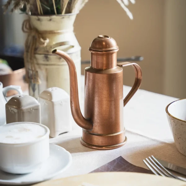 Caffettiera in rame su tavola apparecchiata con tazza di cappuccino, saliera e centrotavola floreale.