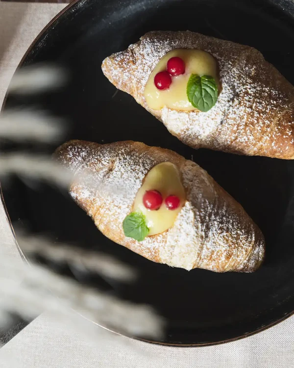 Due croissant su un piatto nero, decorati con crema, frutti rossi e foglie di menta.