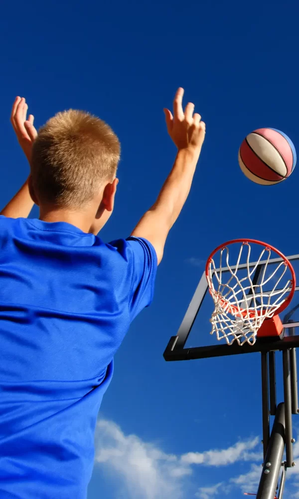Ein Junge im blauen T-Shirt wirft einen Basketball in Richtung Korb unter klarem Himmel.